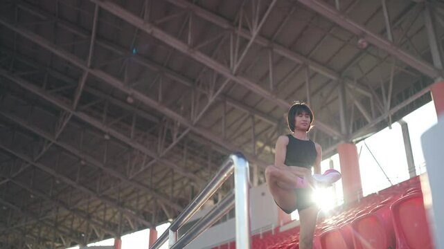 A woman is standing on a bleacher in a stadium. She is wearing a black tank top and she is stretching. The stadium is empty, and the sun is shining brightly, creating a warm and inviting atmosphere