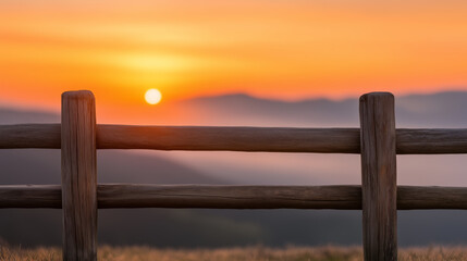 Fototapeta premium A rustic wooden fence against the backdrop of a glowing orange sunset, with soft misty mountains fading into the distance