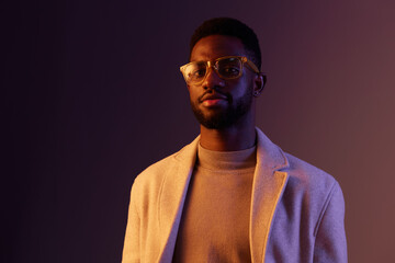Obraz premium Stylish young man in glasses with a neutral expression, wearing a turtleneck sweater and a light overcoat, set against a colorful gradient background
