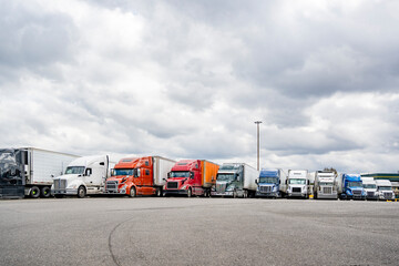 Colorful different big rig semi trucks with semi trailers standing in row on truck stop parking lot...