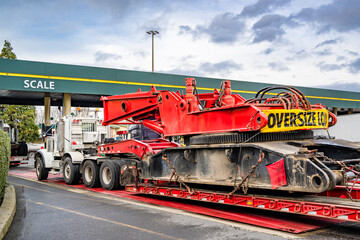White big rig semi truck with oversize load on the step down heavy duty semi trailer going to the scale for weighing the load on the axles for safe driving on highways