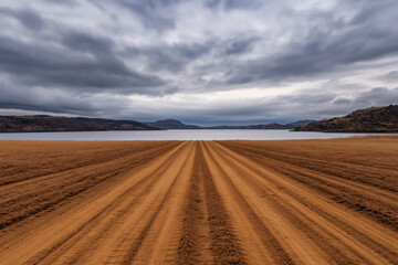 Obraz premium Freshly plowed farmland leading to a calm lake surrounded by hills under a dramatic cloudy sky