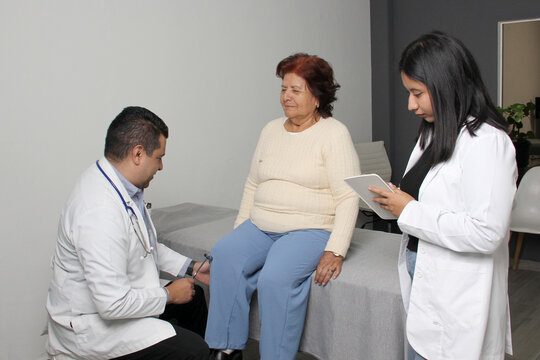 Male orthopedic doctor evaluates the reflexes of his patient's legs with the help of his brunette Latina practitioner in the office
