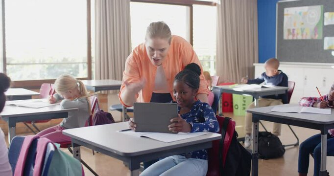 In school, female teacher helping student with tablet in classroom, other students studying