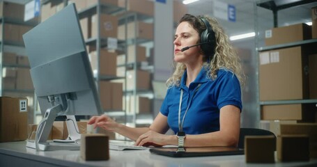 Female customer support specialist in logistics in headset talks to client, uses computer, works in postal service or online store storage. Warehouse employees checking cardboard boxes for delivery.