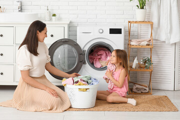 Young woman and her daughter pulling out fresh clothes from washing machine in laundry room