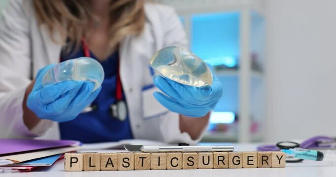 Phrase Plastic Surgery with wooden cubes against woman doctor holding breast implants in hospital. Human body aesthetic alternation concept