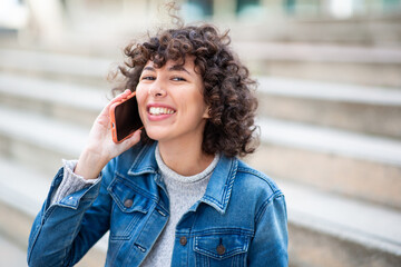 Young woman with curly hair and denim jacket talking on the phone while smiling outdoors