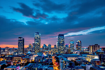Fototapeta premium Urban skyline at dusk with illuminated buildings and dramatic clouds in the background.