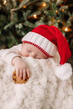 Baby sleeping with Santa hat holding cookie under Christmas tree.