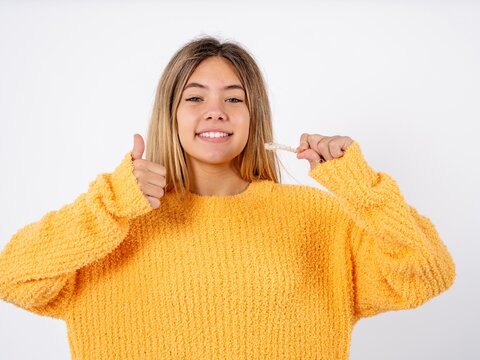 Beautiful caucasian teen girl wearing orange sweater holding an invisible braces aligner and rising thumb up, recommending this new treatment. Dental healthcare concept.
