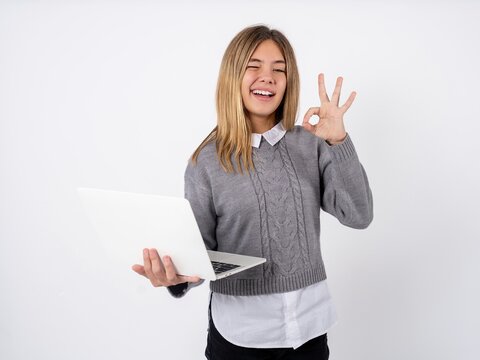 Attractive cheerful skilled Beautiful teen girl wearing grey sweater using laptop showing ok-sign winkin