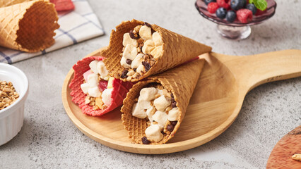 3 ice cream cones with food inside, 2 vanilla cones and 1 red cone.
placed on a wooden tray and has various ingredients including cherries  
All laid out on the table cream color
