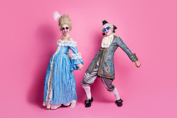 A joyful couple in baroque costumes pose against a vivid pink background, embodying elegance and humor at a lively masquerade.
