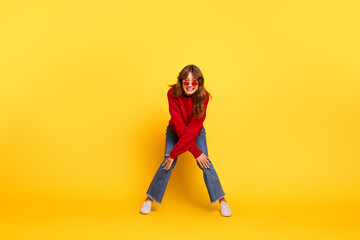 Young woman wearing fashionable red sweater and stylish sunglasses posing on vibrant yellow background, exuding charm and joy
