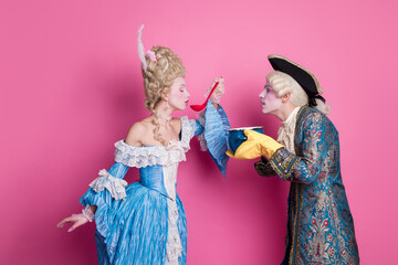 Elegant couple in baroque costumes sharing a playful moment against a pink backdrop, embodying...