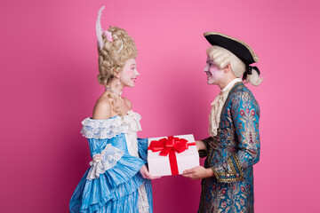Charming couple in extravagant historic costumes exchange a gift against a vibrant pink background