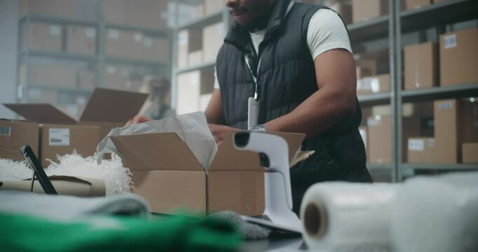Close Up of African American Postal Worker, Shipping Clerk Packing Cardboard Parcel with E-Commerce Order, Scanning Code, Preparing Package for Delivery. Warehouse, Sorting Center of Postal Service.