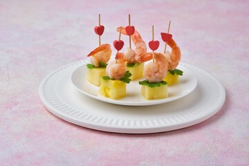 Appetizer of fresh pineapple cubes and shrimps marinated in sauce on a skewer on a white ceramic plate on a pink concrete background. Party snacks.
