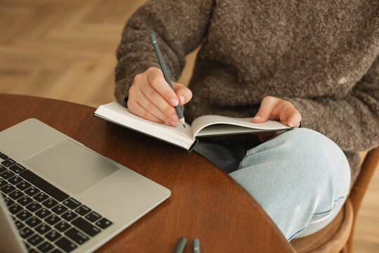 Close-up of a young woman sitting at her dining table using her laptop and writing in a note pad