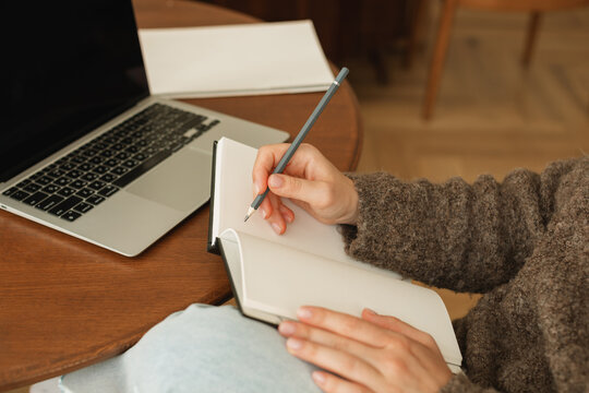 Close-up of a young woman sitting at her dining table using her laptop and writing in a note pad