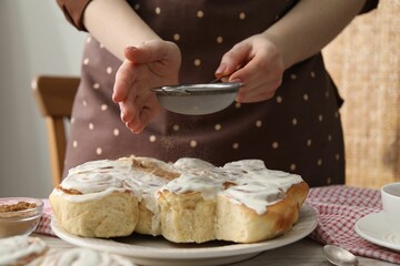 Woman with sieve adding cinnamon powder onto freshly baked rolls at table indoors, closeup