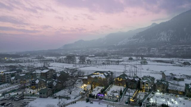 Aerial drone shot flying forward showing lit decorated houses in snow covered pahalgam rafting point valley with himalaya mountains and pink sunset sky in background in Kashmir India