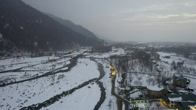Locked aerial drone dusk shot with lit houses, lights from cars passing in the snow covered lidder river valley in pahalgam Kashmir a famous tourist spot