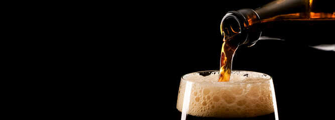 closeup of pouring dark beer from a bottle into a beer pint with foam isolated on black background banner with copy space, delicious drink concept with beautiful lighting