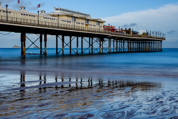 Fototapeta premium Seaside pier with reflections in the sand, long exposure with smooth sea, blue sky and sunshine. Dramatic, Sandy beach holiday resort. 