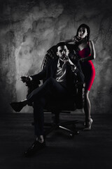 Portrait of rich gentleman in formal wear sitting in leather chair holding glass with cognac and cigar in hands, pretty charming lady in purple dress standing near isolated on grey background