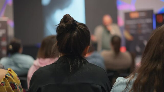 Back of brunette, with her hair in a ponytail, sitting at seminar. Creative minds gather for self-improvement lecture, blurry figure conducts presentation on stage