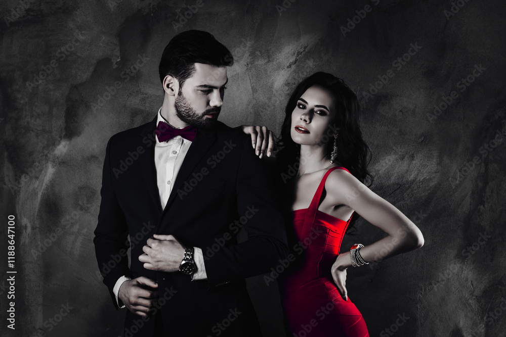 Poster Portrait of virile man with stubble in tuxedo with bowtie correcting fixing buttons on his jacket looking at his charming tempting rich wealthy lover with hairstyle curls isolated on grey background