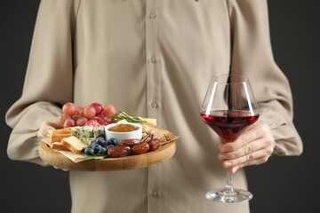 Woman holding board with different types of delicious cheese, other snacks and wine on grey background, closeup