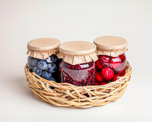Wicker basket holding three jars of preserved berries, blueberries, cranberries, and cherries sealed with paper lids on neutral background. Concept of homemade, natural, healthy and artisanal food
