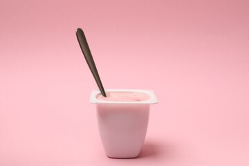 Delicious yogurt in plastic cup with spoon on pink background