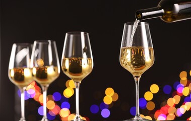 Pouring white wine into glass against black background with blurred lights, closeup. Bokeh effect