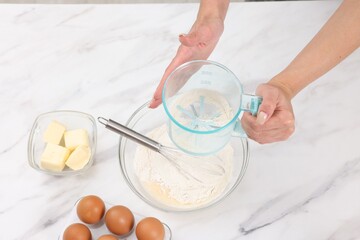Woman adding flour into bowl with dough at white marble table, above view