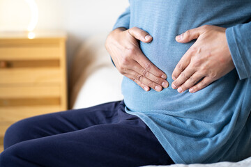 Man sitting alone on his bed and suffering from stomach cramps while home alone.Abdominal pain can...