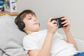 A ten-year-old boy calmly plays on a smartphone while lying on a bed with headphones.