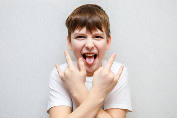 cute ten year old boy looking at camera standing against wall and making rock gesture with crossed...