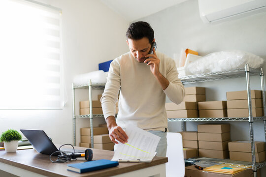 Latin entrepreneur managing orders and logistics in his small business or sme, checking paperwork and talking on the phone