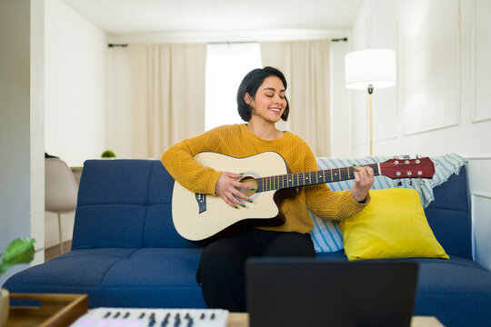 Hispanic musician sitting on sofa, strumming acoustic guitar during home recording session or online music lesson - Powered by Adobe