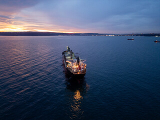 Illuminated cargo ship bulk carrier anchored on the calm sea at twilight, with city lights visible...