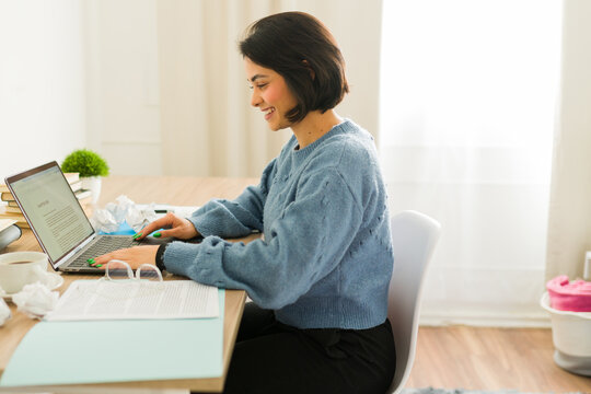 Profile view of a smiling female author working on her new novel, typing on a laptop and surrounded by crumpled paper and books