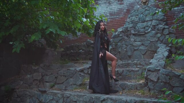 Fantasy woman Medusa gorgon Greek goddess. Black gothic carnival costume. Girl Fashion model posing in outdoor. On head snakes scary hair, black silk hood long dress, cape fly in motion. Dark queen