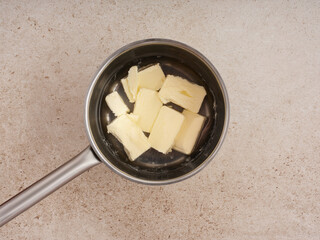 A saucepan holds several cubes of butter as they slowly melt on the stovetop. The kitchen countertop is light-colored, indicating a tidy cooking area