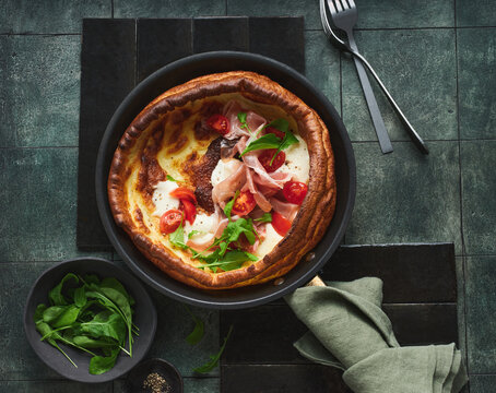 A delectable Yorkshire pudding is filled with slices of prosciutto, creamy mozzarella, and vibrant cherry tomatoes, garnished with fresh greens, placed on a dark textured surface