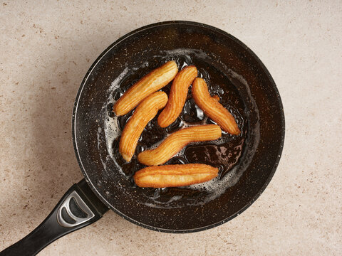 Churros are frying in a pan filled with bubbling hot oil, creating a golden-brown color. This occurs in a cozy kitchen during a bright afternoon, perfect for enjoying freshly made treats