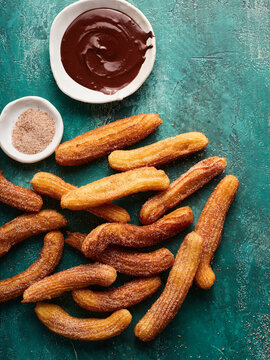 Warm churros are arranged atop a teal surface, accompanied by bowls of rich chocolate sauce and sweet cinnamon sugar. This delightful spread invites indulgence